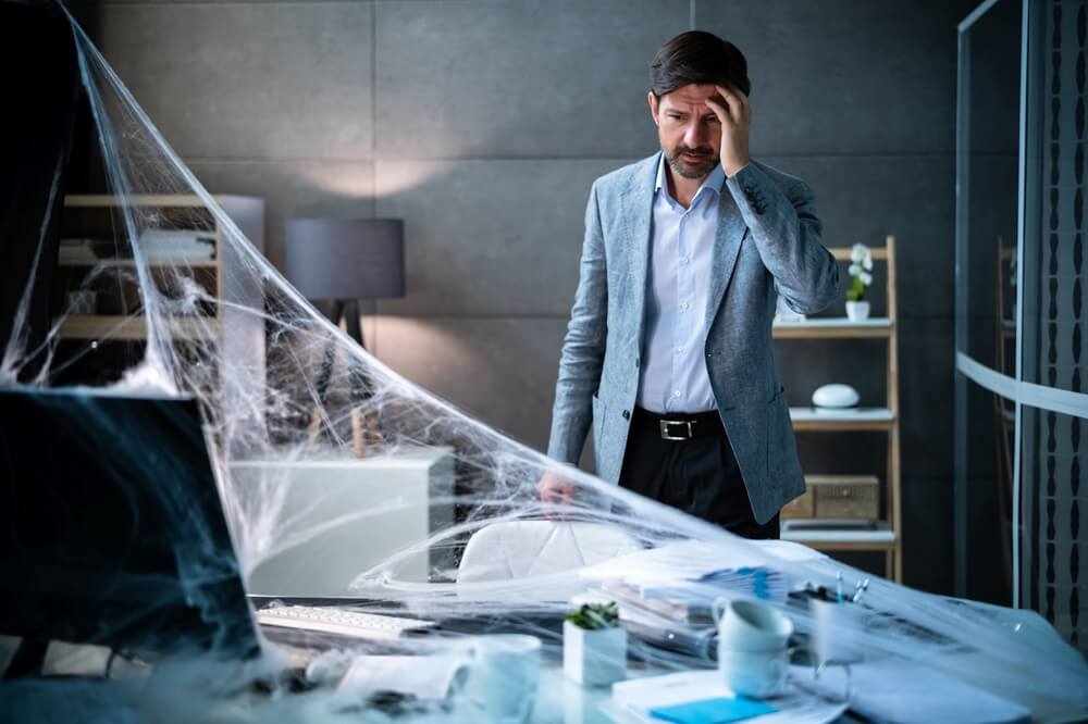 man looking at desk in office covered in cobwebs